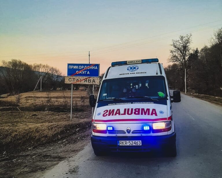 Ambulans z włączonymi sygnałami świetlnymi na drodze przy tablicy strefy przygranicznej