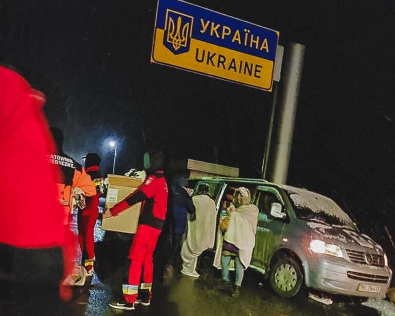 Ratownicy przenoszą kartony nocą pod tablicą Ukraina, obok osoby okryte kocami przy samochodzie